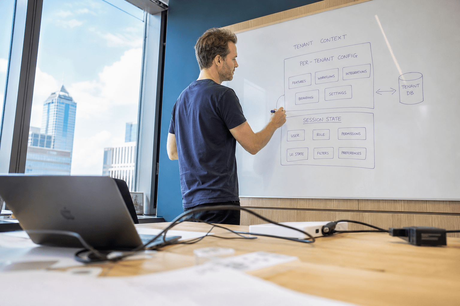 Low-angle view from a desk looking up at an engineer drawing a tenant configuration diagram on a whiteboard in a bright, sunlit office.