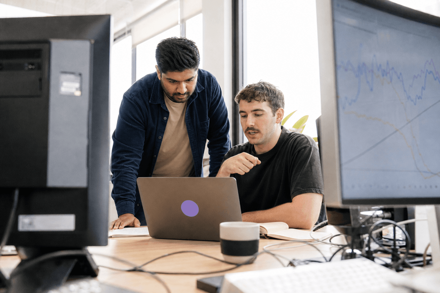 Daylit office scene framed between two monitors, showing two engineers collaborating around a laptop and screen displaying line-chart metrics, with cables and a coffee cup on the desk.