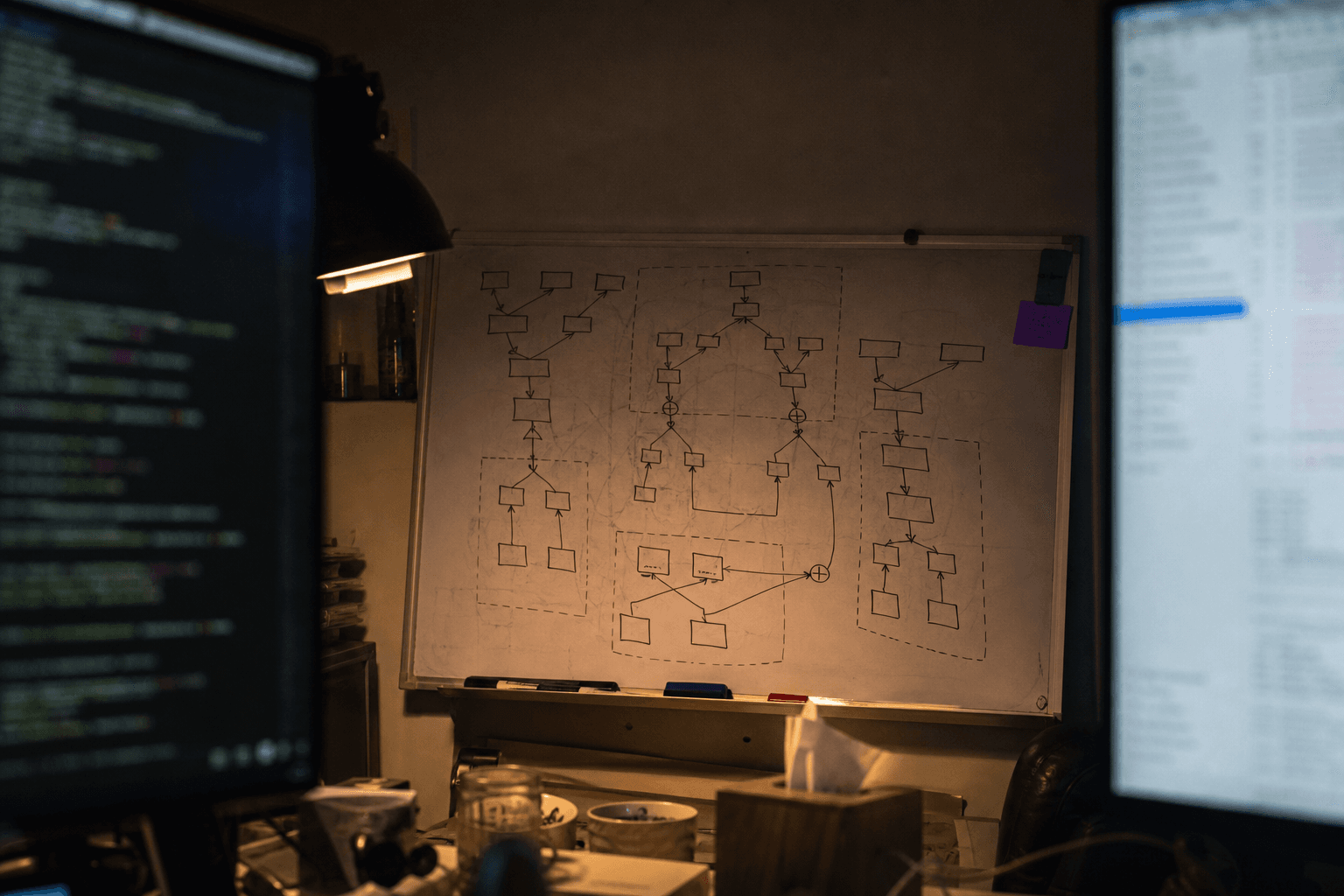 View framed between two monitor bezels looking toward a whiteboard covered in hand-drawn computational graph diagrams with operator fusion annotations, lit by warm task lamp glow in a dark engineering workspace.