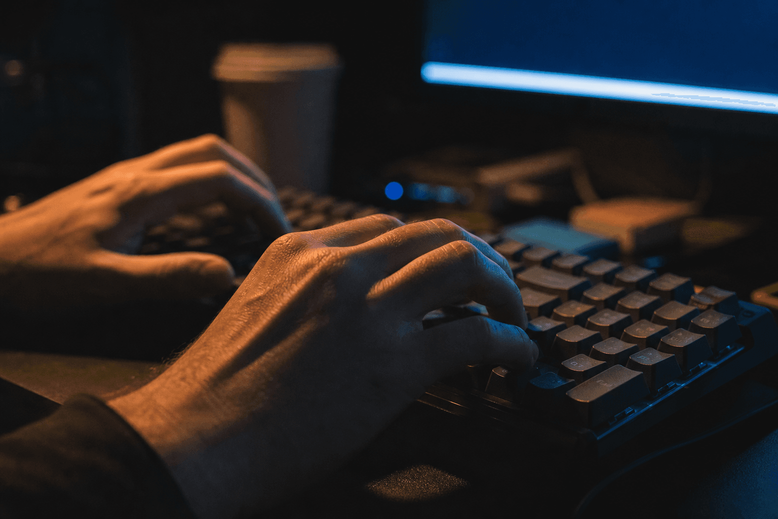 A developer's hands rest on a mechanical keyboard in a darkened office workspace, lit by the cool glow of a monitor and a warm amber desk lamp, with blurred desk accessories visible in the background.