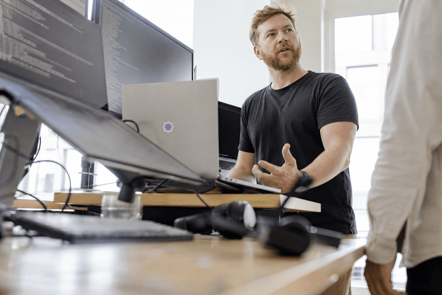 Low-angle view past keyboards and monitors toward an engineer standing at a desk in a bright, daylight-filled office discussing something with a colleague.