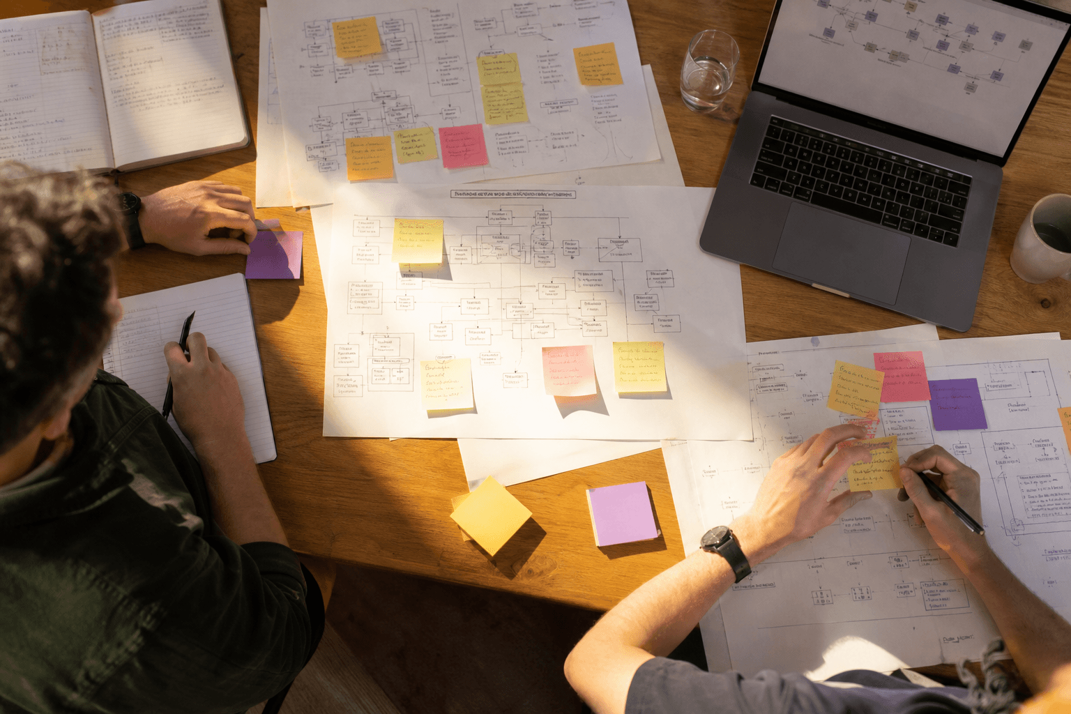 Overhead view of a desk covered in printed system diagrams and coloured sticky notes, with two people's hands sorting the papers in warm afternoon light.